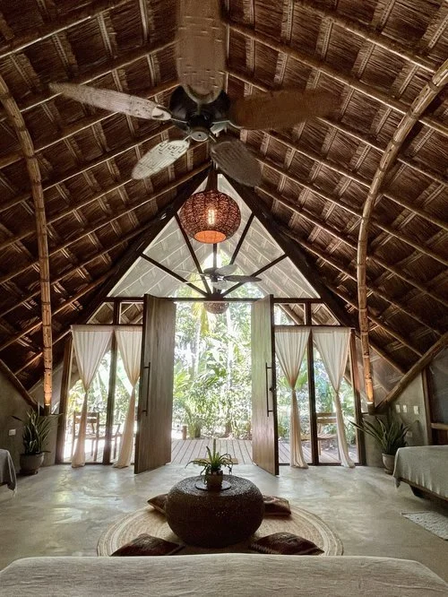 Interior view of a tropical thatched roof room with open double doors leading to lush greenery outside, decorated with potted plants, a hanging lamp, ceiling fans, and simple furniture.