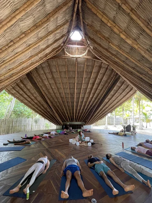 People practicing yoga or meditation in a large open-air, thatched-roof pavilion with a wooden floor, surrounded by lush greenery.
