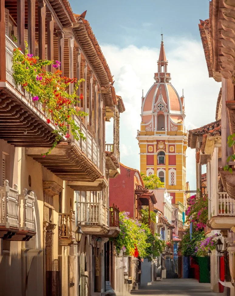 Colorful street with European-style buildings, balconies with flower boxes, and a tall ornate church tower in the background under a partly cloudy sky.