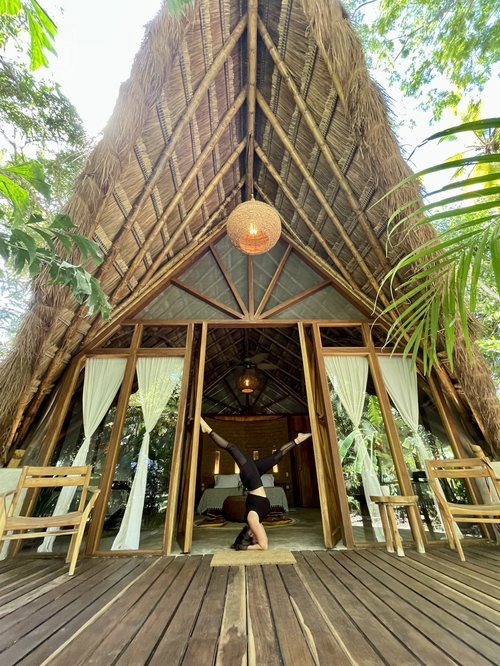 Person doing a headstand in front of a Tiki-style treehouse with wooden deck, chairs, and white curtains, surrounded by green jungle plants.