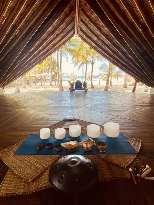View from inside a large open-front hut with a wooden floor, overlooking a tropical beach with palm trees and ocean in the background. Inside, there's a mat with candles, seashells, and percussion instruments for a meditation or sound bath session.
