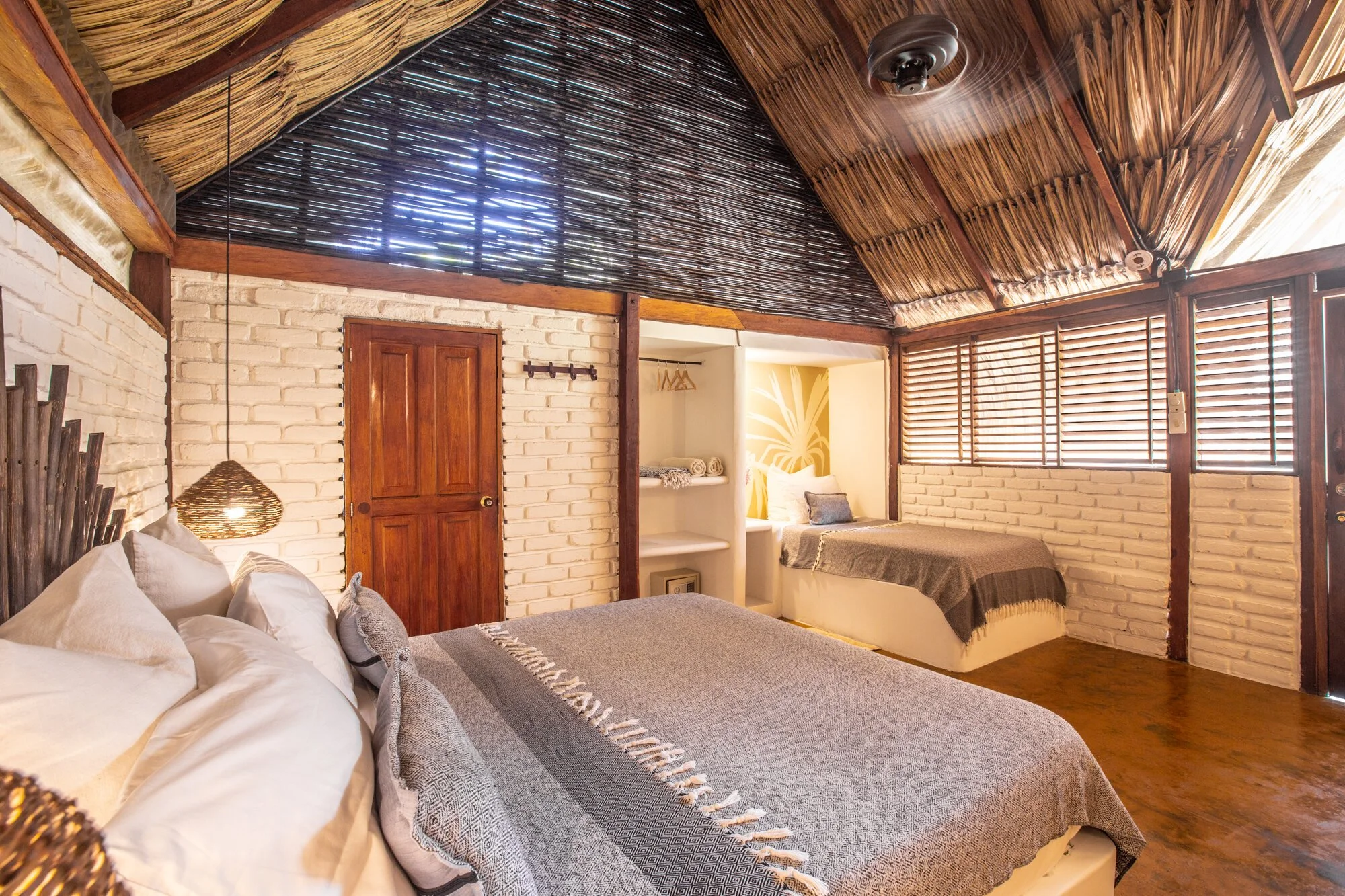 A rustic bedroom with a white brick wall, wooden ceiling and blinds, featuring a large bed with white bedding and multiple pillows, a small pendant light, and a built-in niche with a bed and decorative wall art.