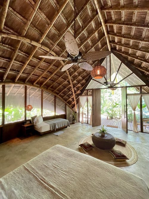 Interior of a tropical-style bedroom with a thatched roof, ceiling fans, a bed, a circular rug with cushions, and large glass doors leading to an outdoor deck surrounded by greenery.