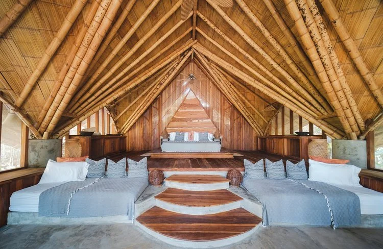 A cozy wooden bedroom with two beds, built into a A-frame bamboo structure with a high vaulted ceiling, natural lighting, and a small stage area at the back.