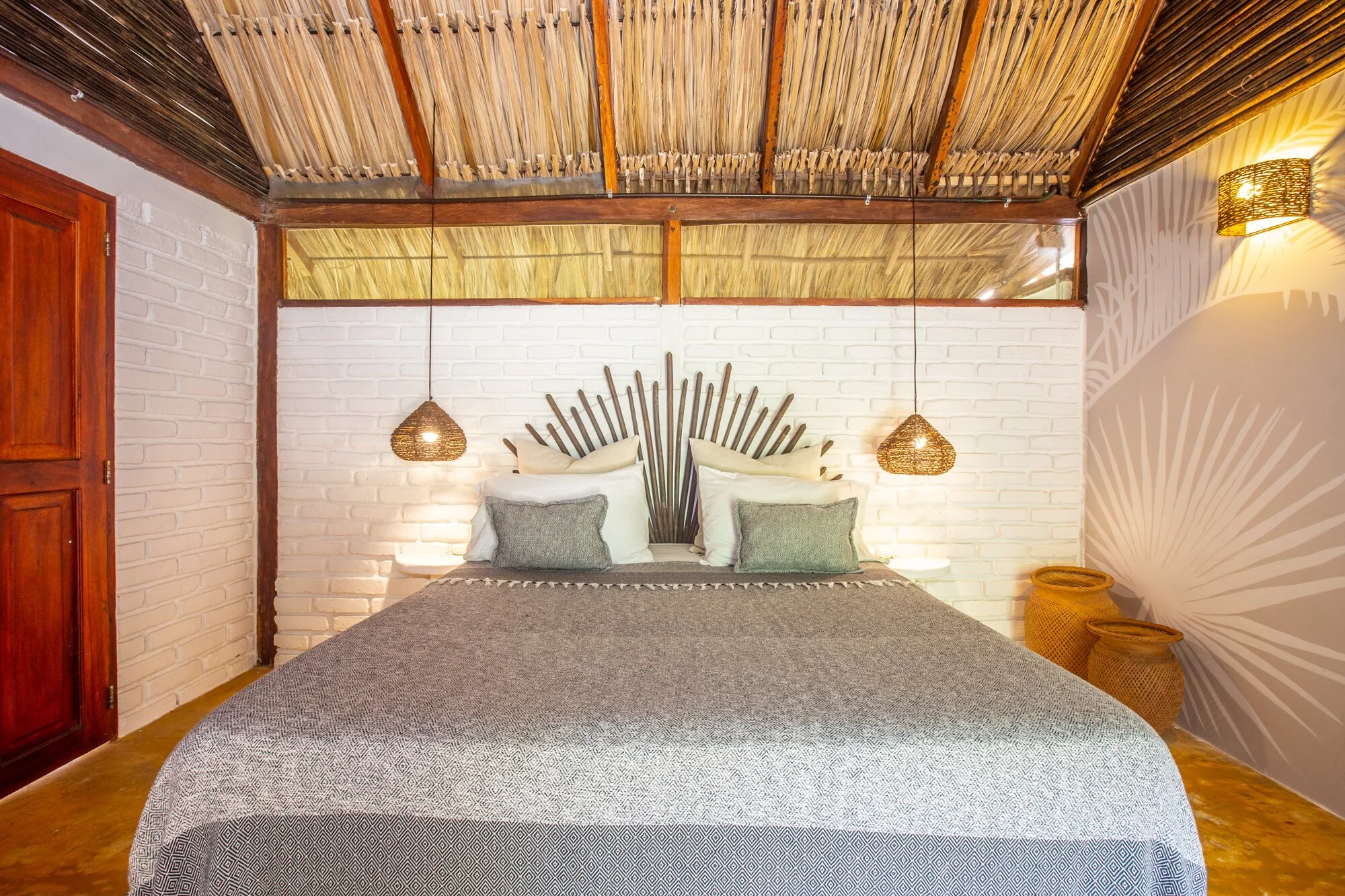 A cozy bedroom with a large bed, white brick wall, and unique headboard design. Two hanging wicker pendant lights flank the bed. The ceiling is made of natural materials, and two woven baskets are placed on the floor beside the bed.