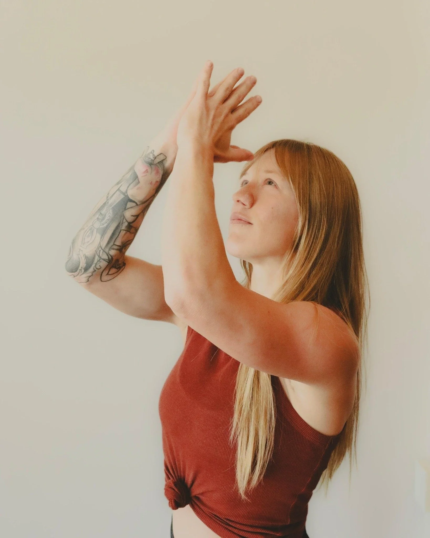 A young woman with long red hair, wearing a sleeveless reddish-brown top, poses against a plain light-colored wall. She has a tattoo sleeve on her left arm and is raising her arms with her hands together, looking slightly upward with a serene expression.