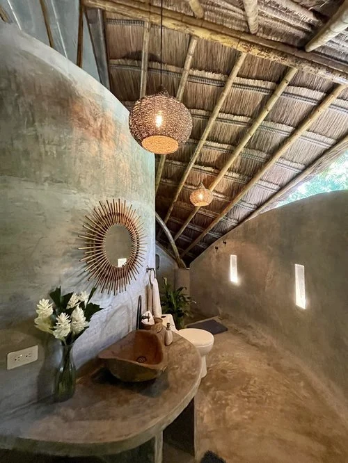 Interior of a rustic bathroom with a curved concrete wall, wooden ceiling, and small windows, decorated with a round mirror, a vase with white flowers, and a stone sink.