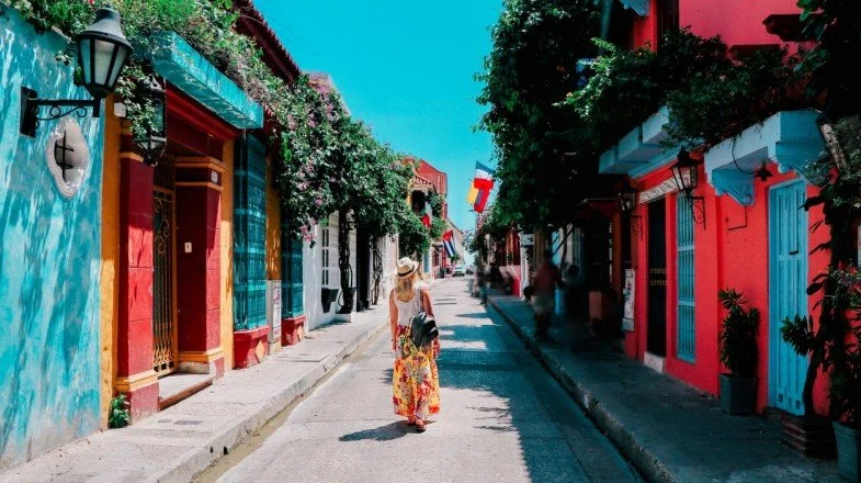 Woman in floral dress and hat walking down colorful street with vibrant buildings and flags.