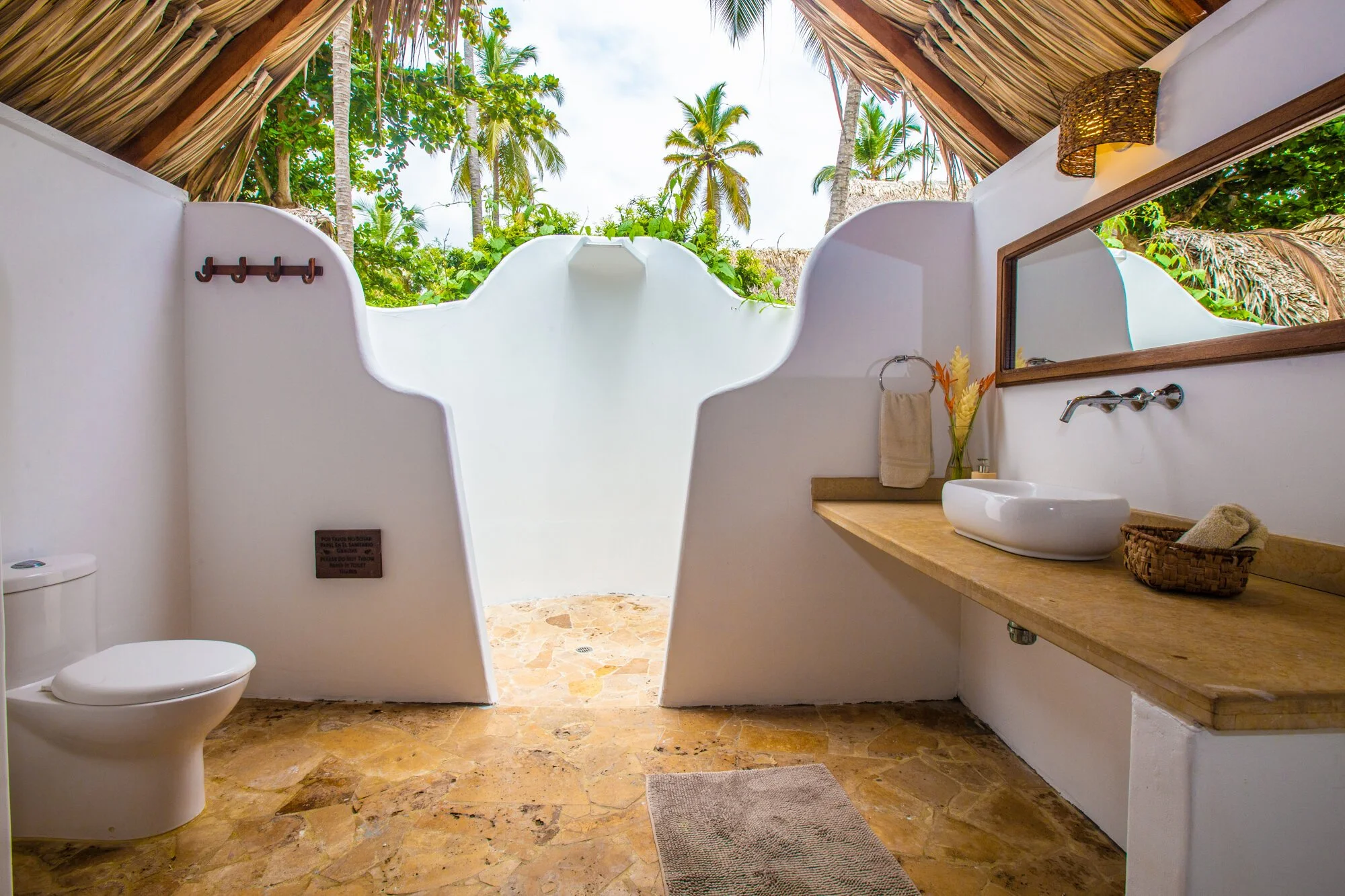Open-air bathroom with a toilet, a wooden countertop with a white basin sink, a mirror, a towel, a basket, and tropical plants outside. The ceiling is made of thatch, and the floor is stone.