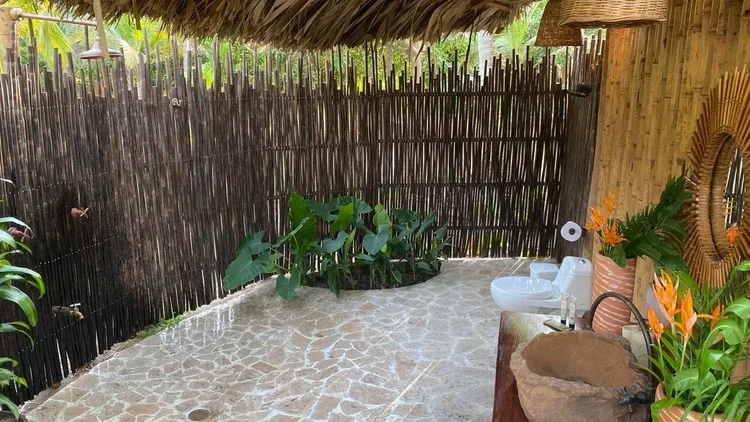 A tropical outdoor bathroom with a thatched roof, bamboo fencing, stone flooring, and lush green plants, including a sink area with a carved stone basin and a mirror surrounded by orange flowers.