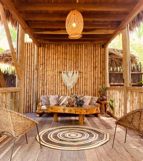 Beachside wooden porch with woven hanging lamp, comfortable sofa with patterned pillows, round wooden table, decorative wall hanging, potted plant, and two wicker chairs on a circular rug.