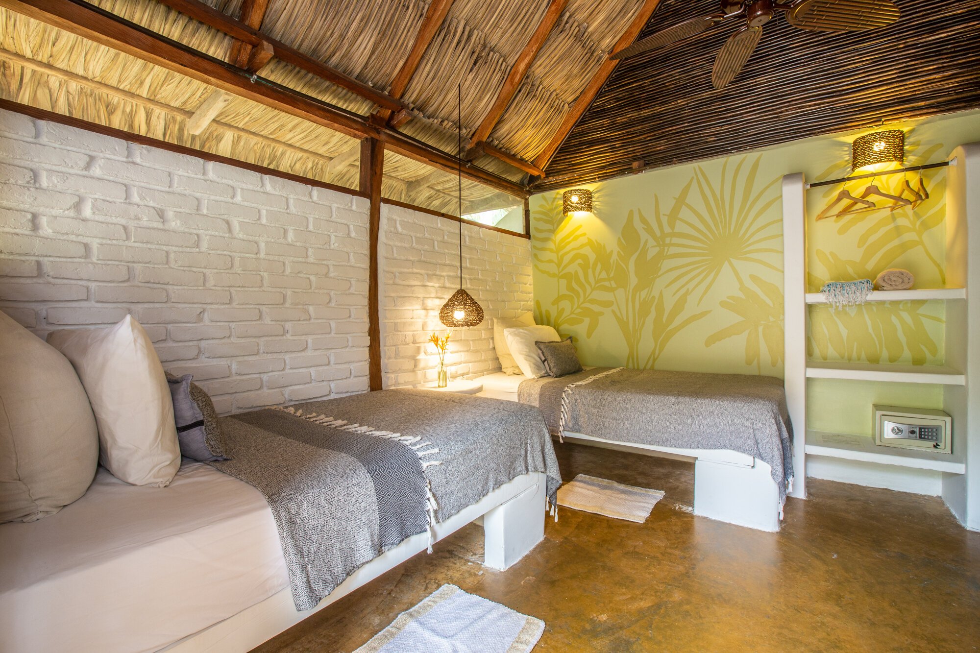 A bedroom with two single beds, gray bedspreads, and pillows. The wall behind one bed has a green tropical mural, and the other wall features a white brick wall. There are hanging wicker lamps, a small bedside table with a lamp, and shelves with towels. The ceiling is made of wood and thatch, and the floor is polished concrete.