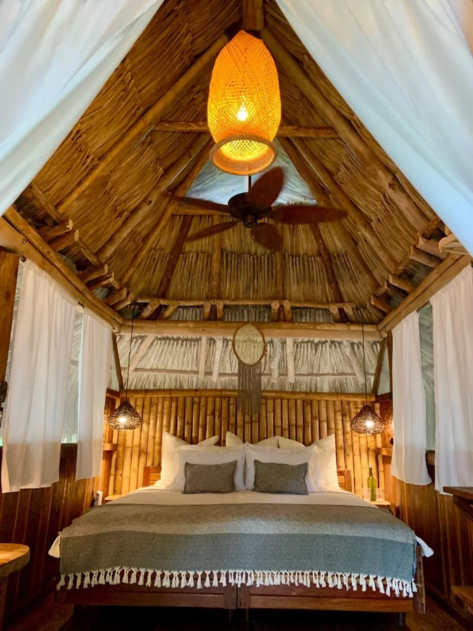 A cozy bedroom with a bamboo and thatched ceiling, white curtains, a wooden bed with white bedding and gray pillows, a dreamcatcher hanging above the headboard, and a ceiling fan with a woven light fixture.