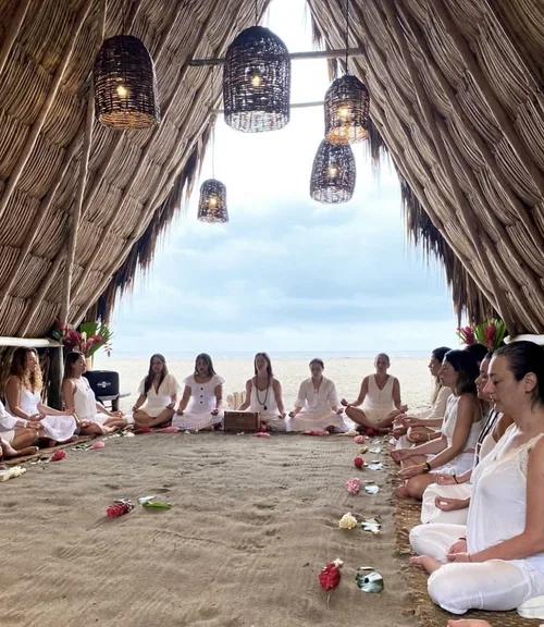 People practicing meditation inside a beach hut with a thatched roof and hanging lanterns, facing the ocean.