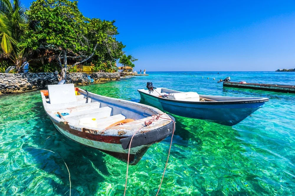 Two small boats floating in clear turquoise water near a tropical shoreline with lush green trees and a rocky wall, under a bright blue sky.