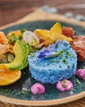 Colorful edible arrangement with blue rice, fruits, vegetables, and decorative flowers on a dark plate.