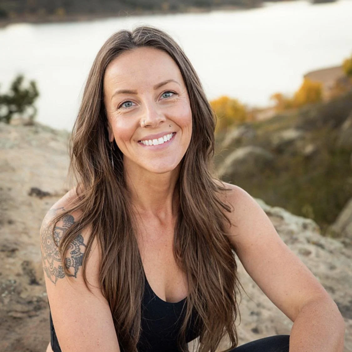 A smiling woman with long brown hair, tattoos on her left shoulder, wearing a black top, outdoors near rocks and trees during sunset or sunrise.