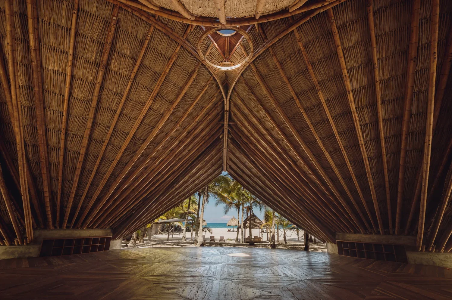 View from inside a thatched beach hut with an open front, showing a sandy beach, palm trees, and the ocean in the distance.