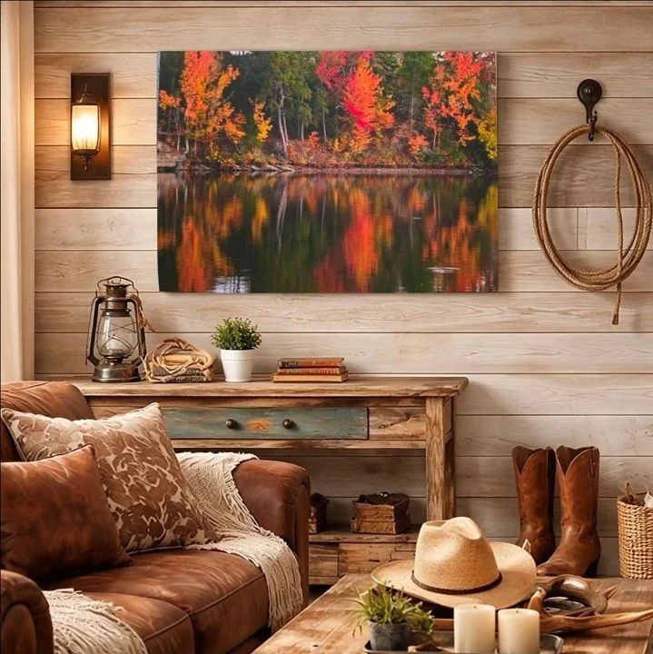 A portrait of autumn trees on a lake hangs on a wooden wall over a living room with wooden tables and a brown leather couch. Cowboy boots, a cowboy hat, books, lanterns, and candles sit in the room.. Soft, inviting sunlight fills the room.