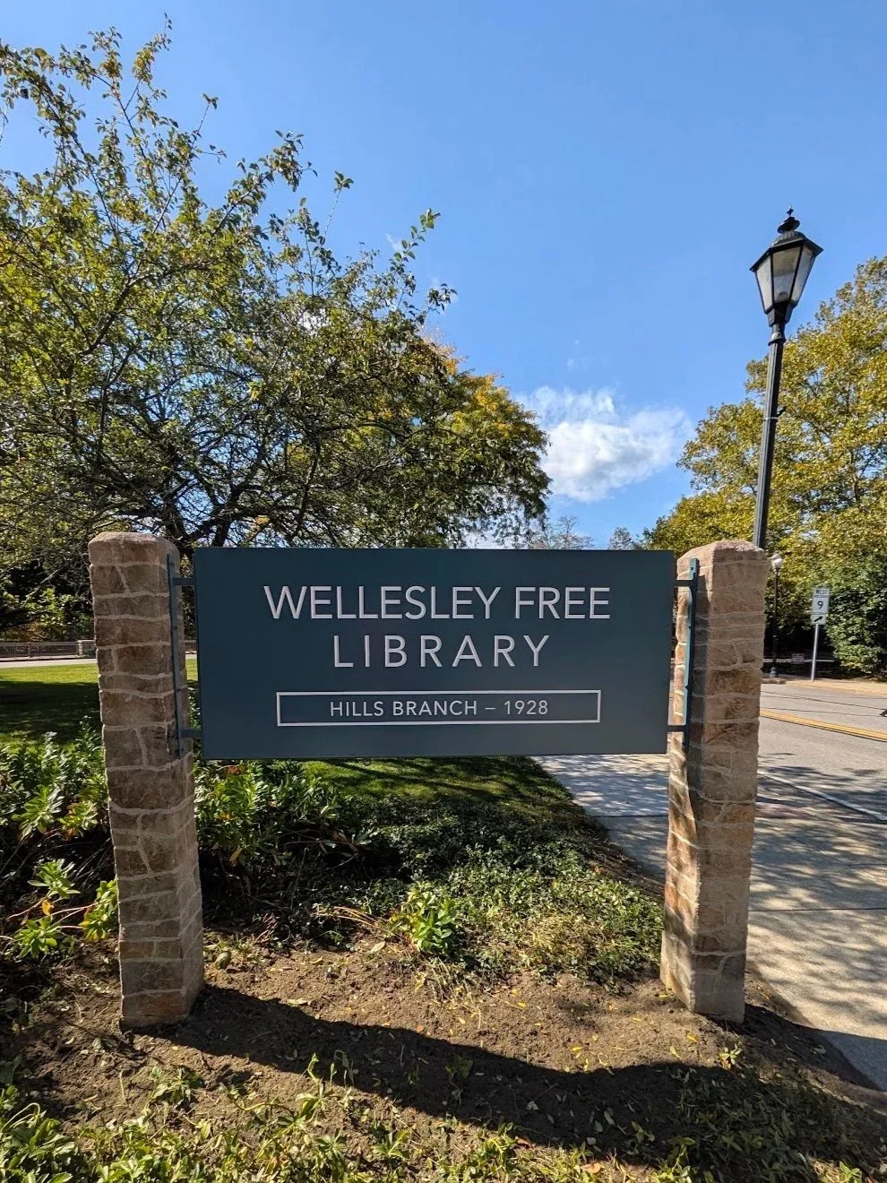 A green wooden sign between stone posts. In white lettering, the sign says: Wellesley Free Library, Hills Branch - 1928. The sign is on a tree-lined street.