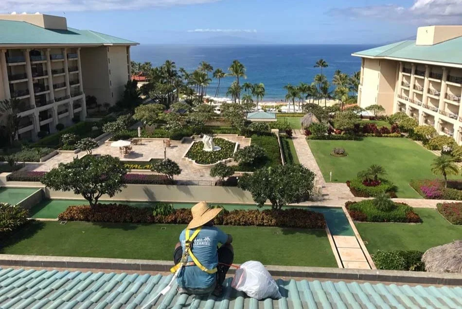 Worker in a straw hat and safety harness on a resort's tile roof edge, overlooking gardens and the Maui coast