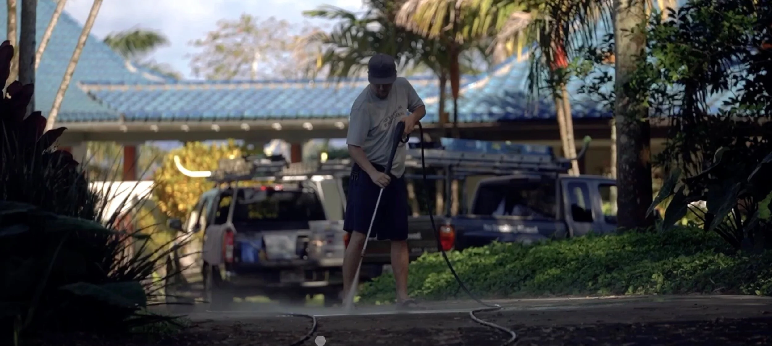 Technician pressure washing a driveway beside work trucks with ladder racks, palm trees behind