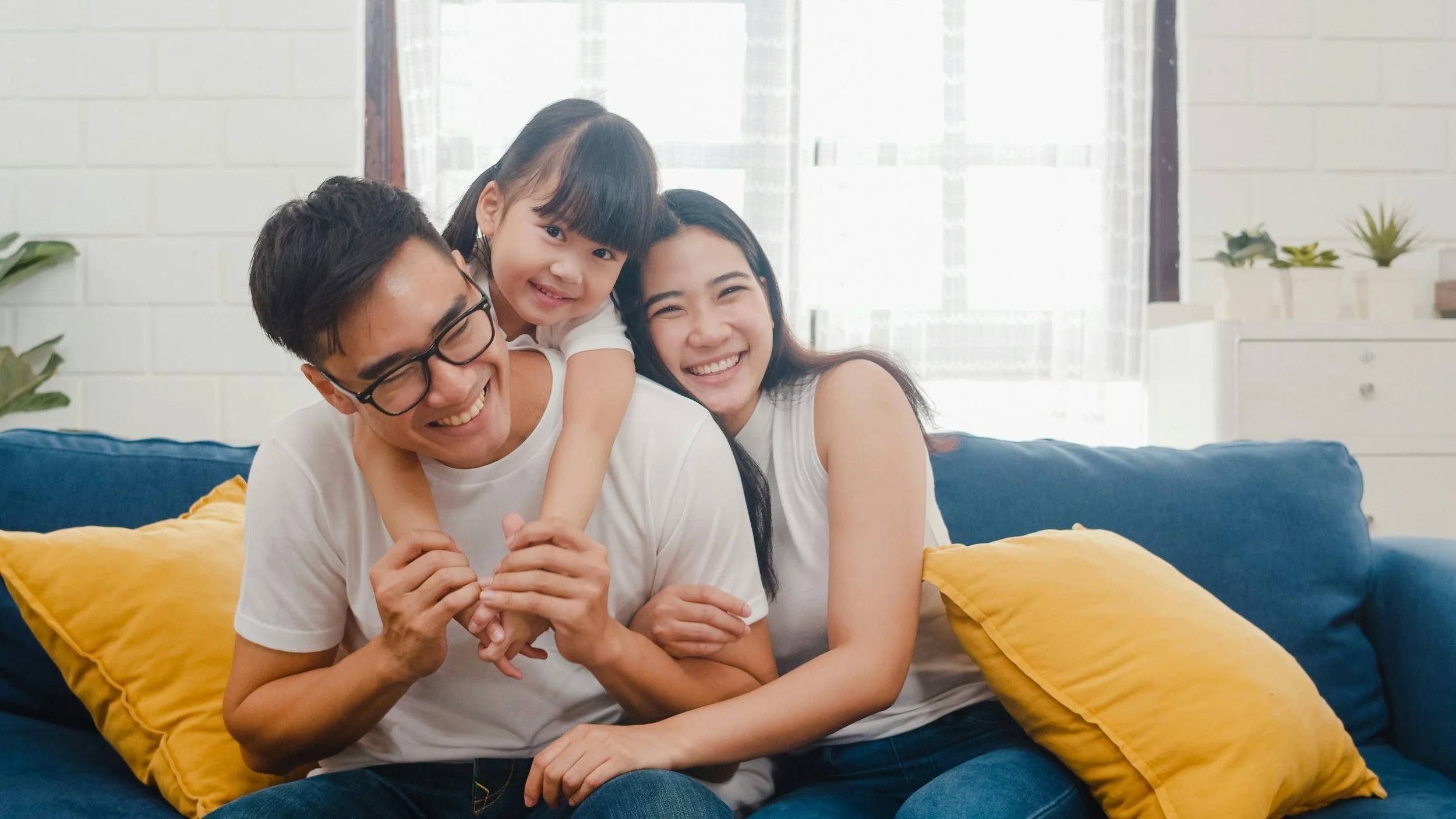 A family of four, two adults and two children, sitting together on a blue couch in a bright living room, smiling and enjoying each other's company.