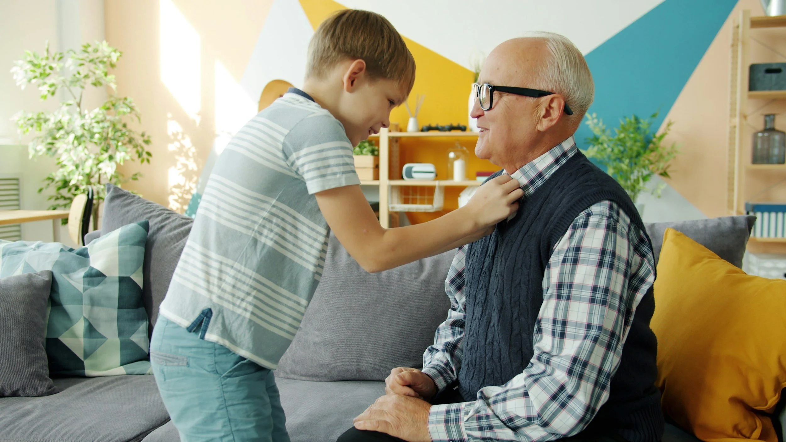 A young boy in a striped shirt and light blue shorts is standing close to an elderly man with white hair, black glasses, and a checkered shirt underneath a navy vest. The boy is playfully grabbing the man's tie, and they are smiling and engaging in a lighthearted moment in a brightly lit living room with colorful wall art and green plants.
