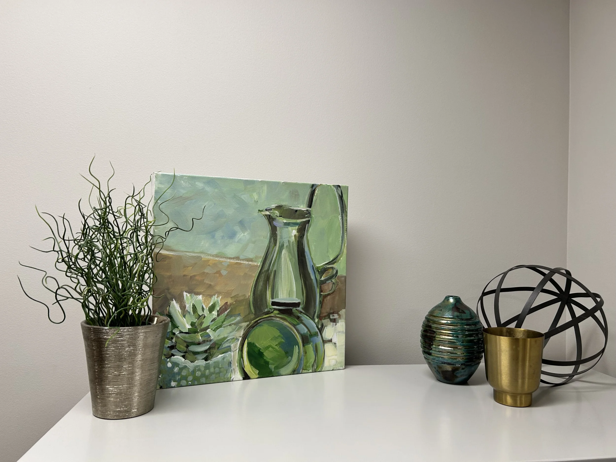 Decorative table display with potted plant, abstract painting, glass vase, striped ceramic vase, gold cup, and wireframe chair against a plain white wall.