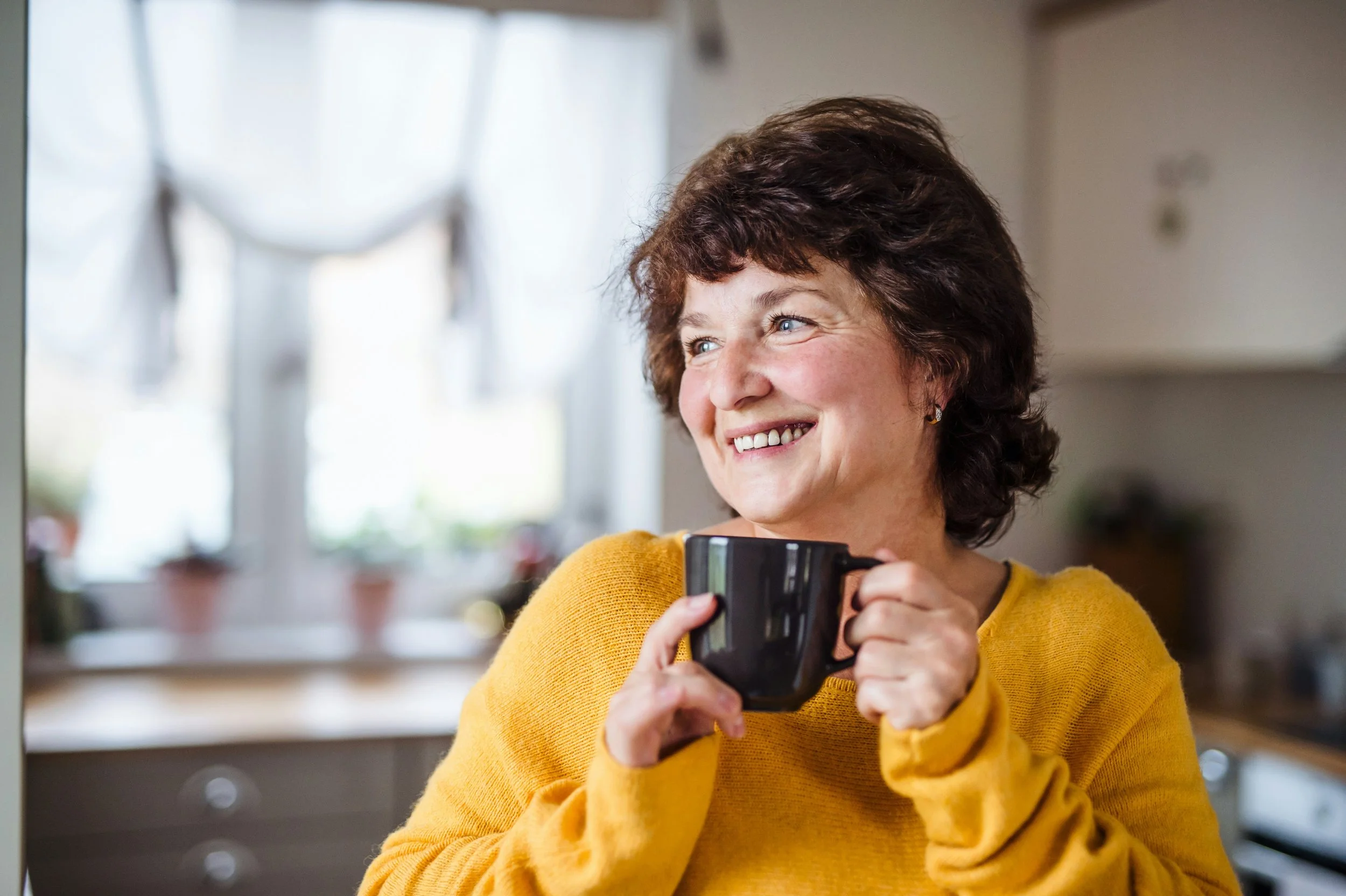 A woman with short curly brown hair and blue eyes smiling while holding a camera in a kitchen with bright windows.