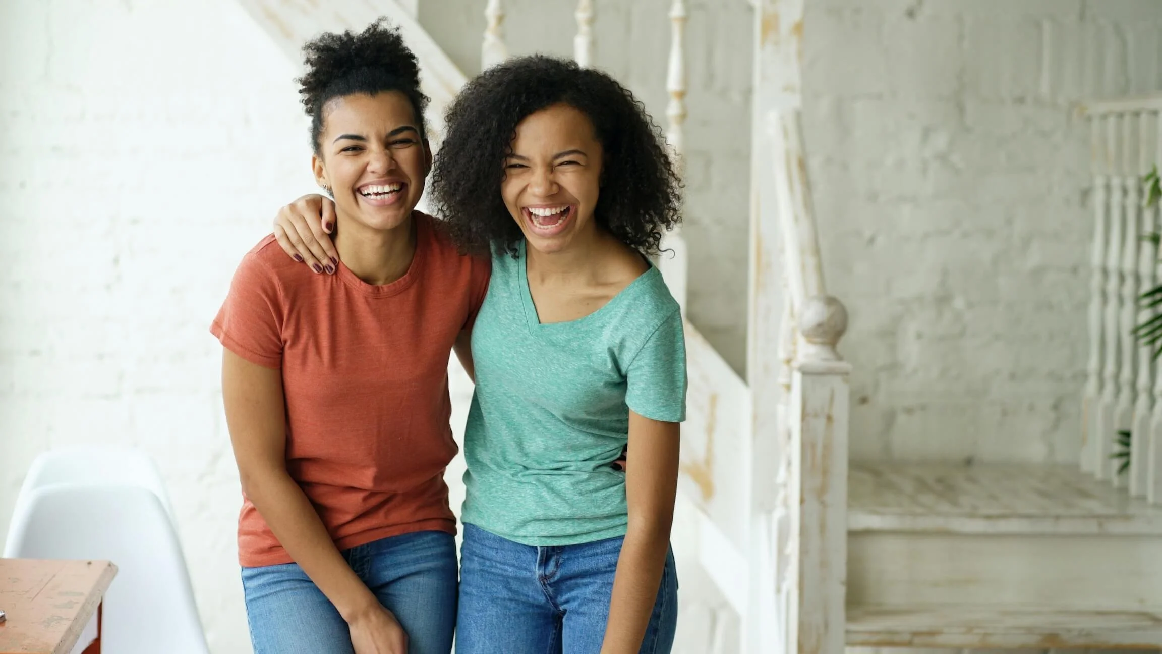 Two women laughing and hugging each other indoors, with a white brick wall and staircase in the background.