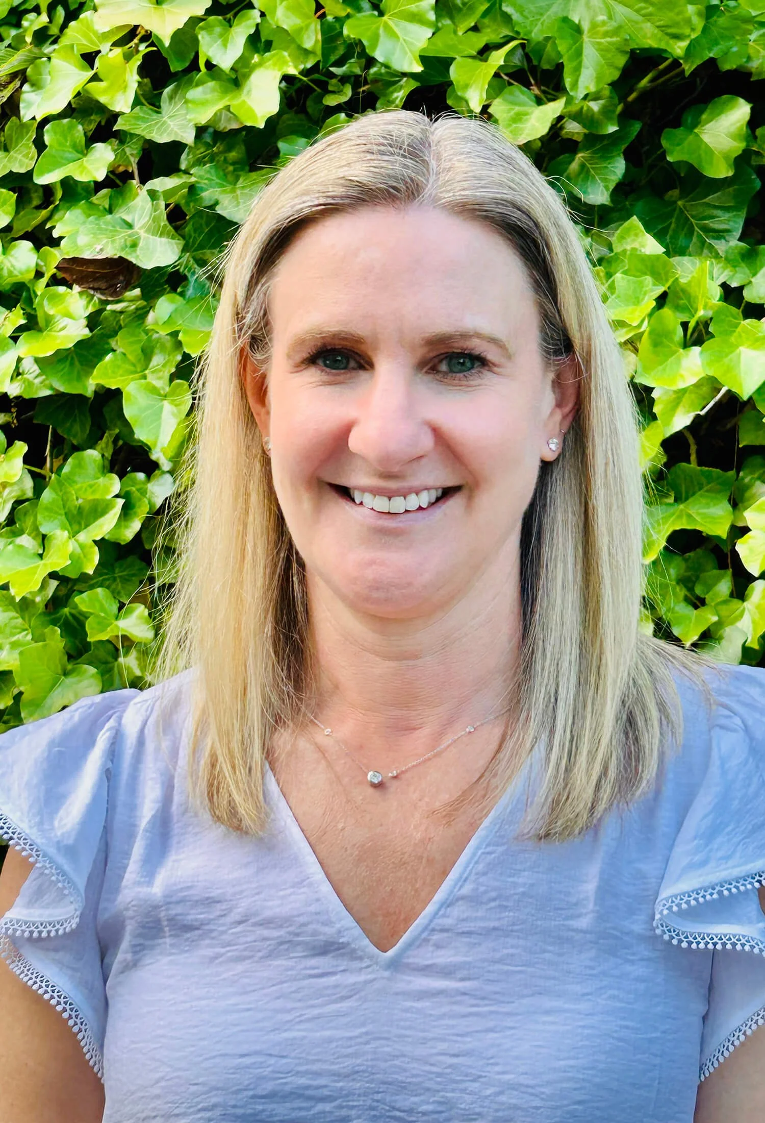A woman with blonde hair, blue eyes, and earrings smiling, wearing a white blouse with lace details, standing outdoors in front of green ivy leaves.