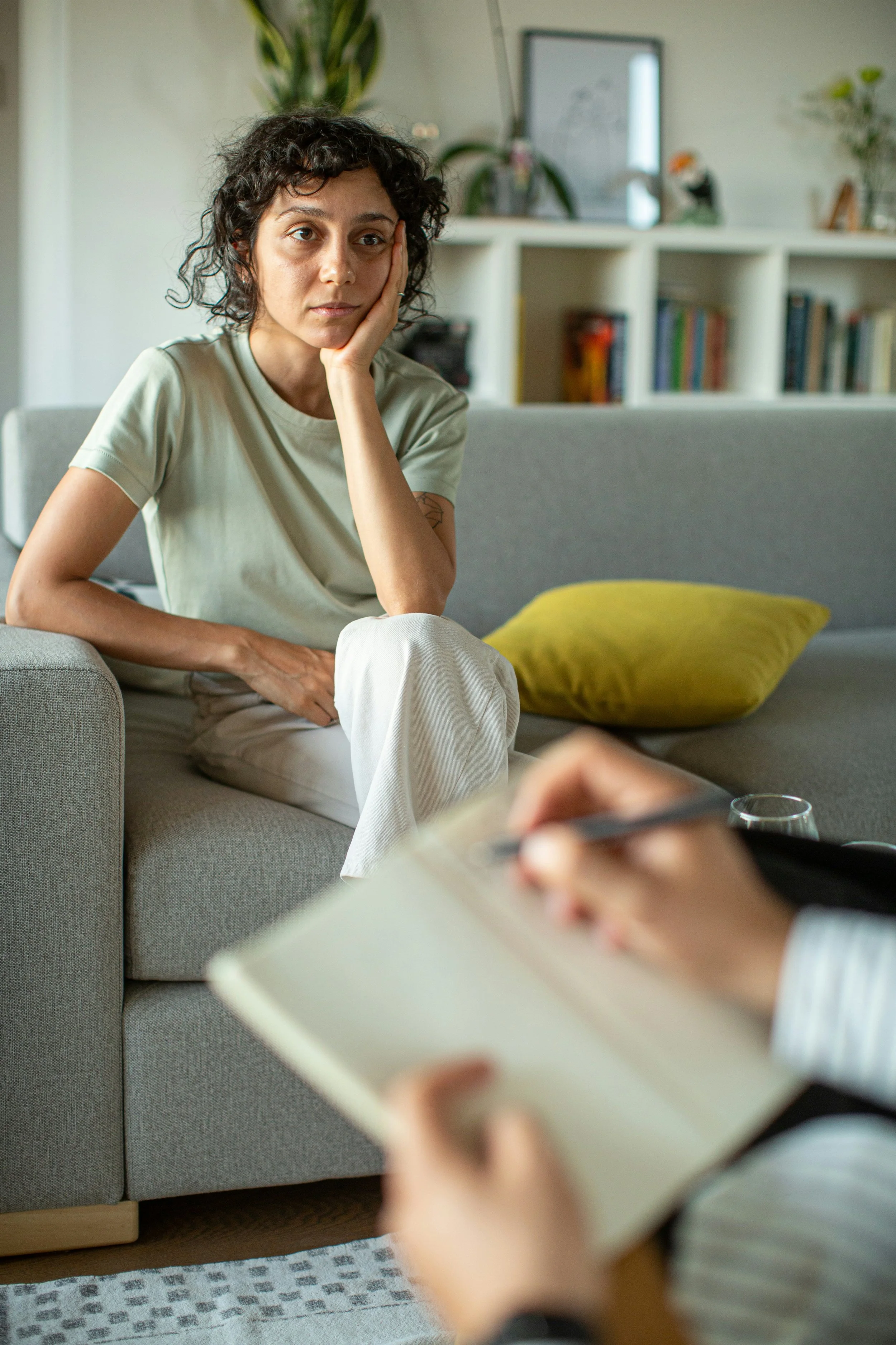 A woman with short curly dark hair, wearing a beige t-shirt, sitting on a gray couch with a yellow pillow. She rests her head on her hand and looks thoughtful or tired. In front of her, someone is taking notes with a pen and paper.