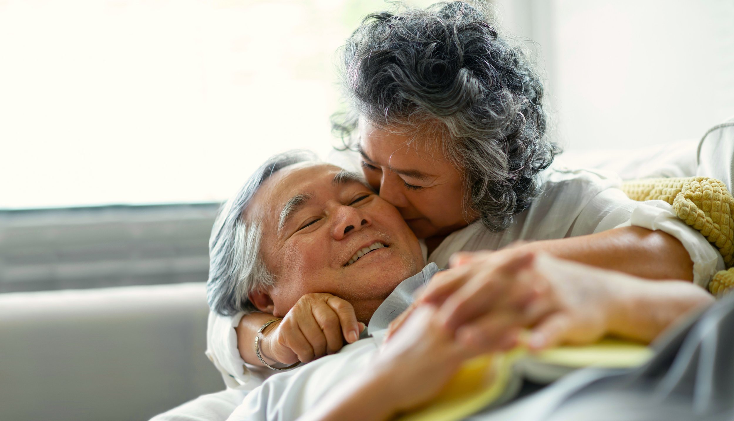 An elderly woman kisses an elderly man on the cheek while lying in bed. They are smiling and appear happy.