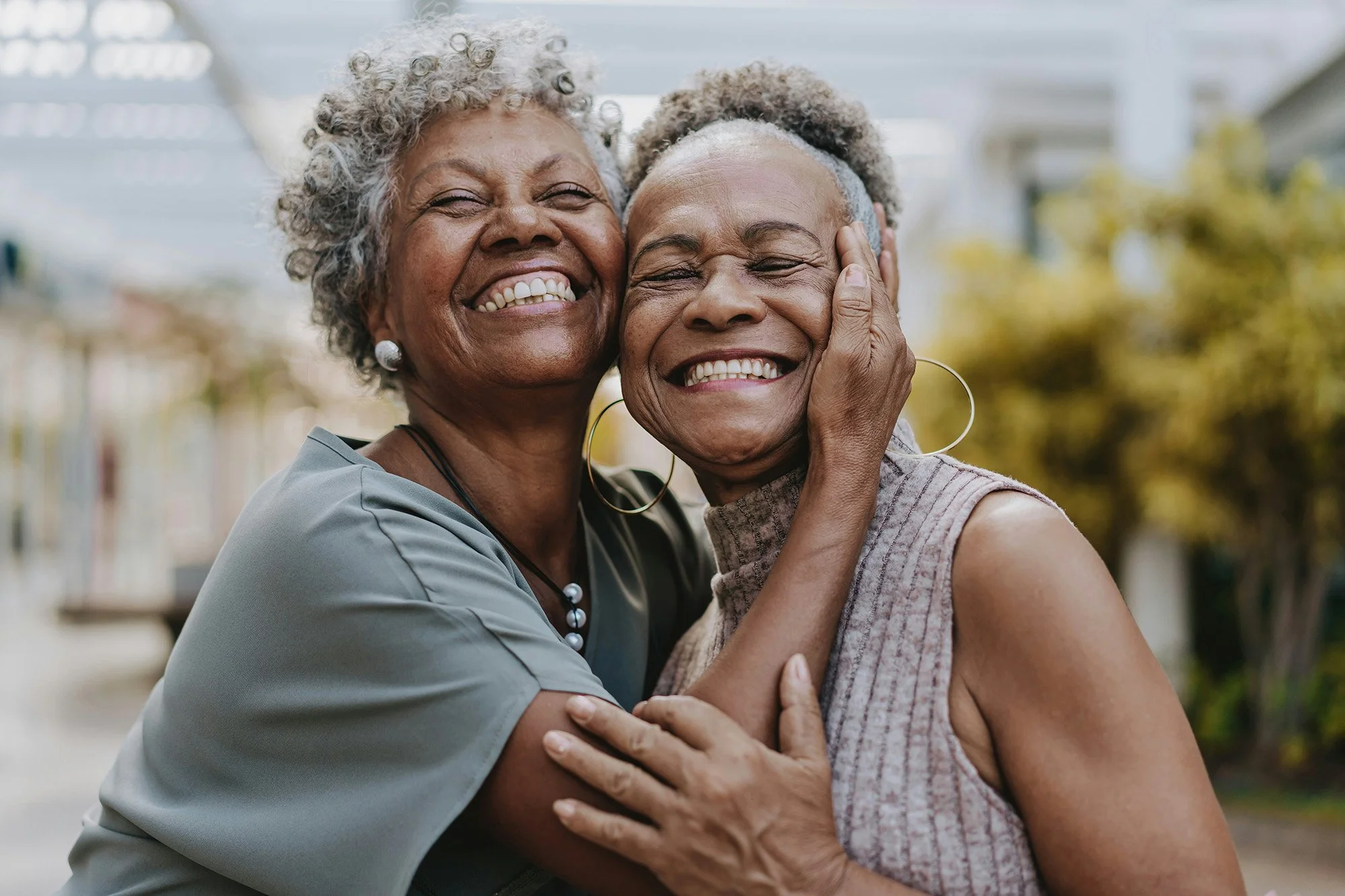 Two elderly women, with gray curly hair, are hugging and smiling joyfully outdoors on a sunny day, with a blurred background of trees and a house.