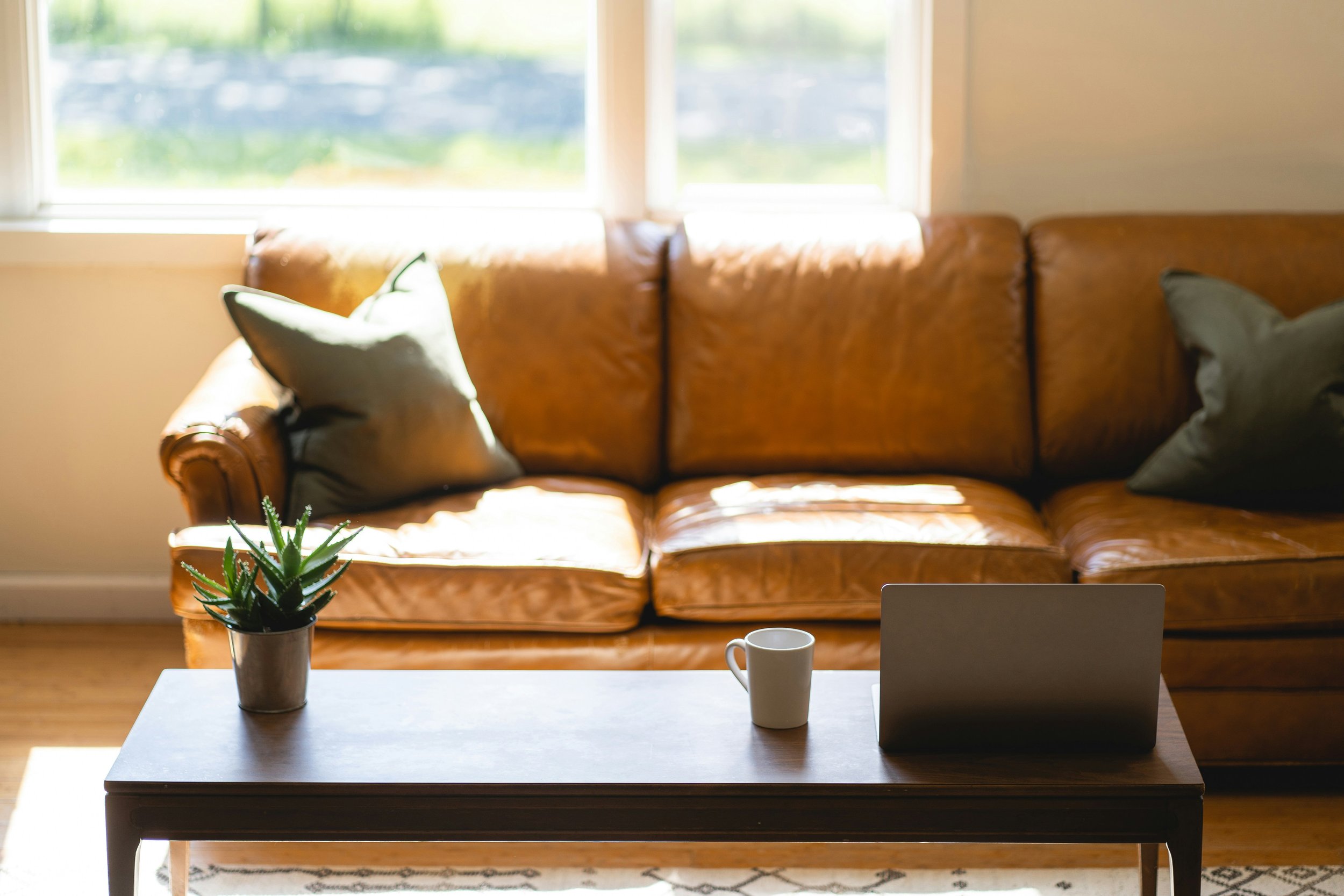 Living room with a brown leather sofa, green and gray pillows, a white coffee table with a potted succulent, a white mug, a laptop, and natural light coming through large windows.