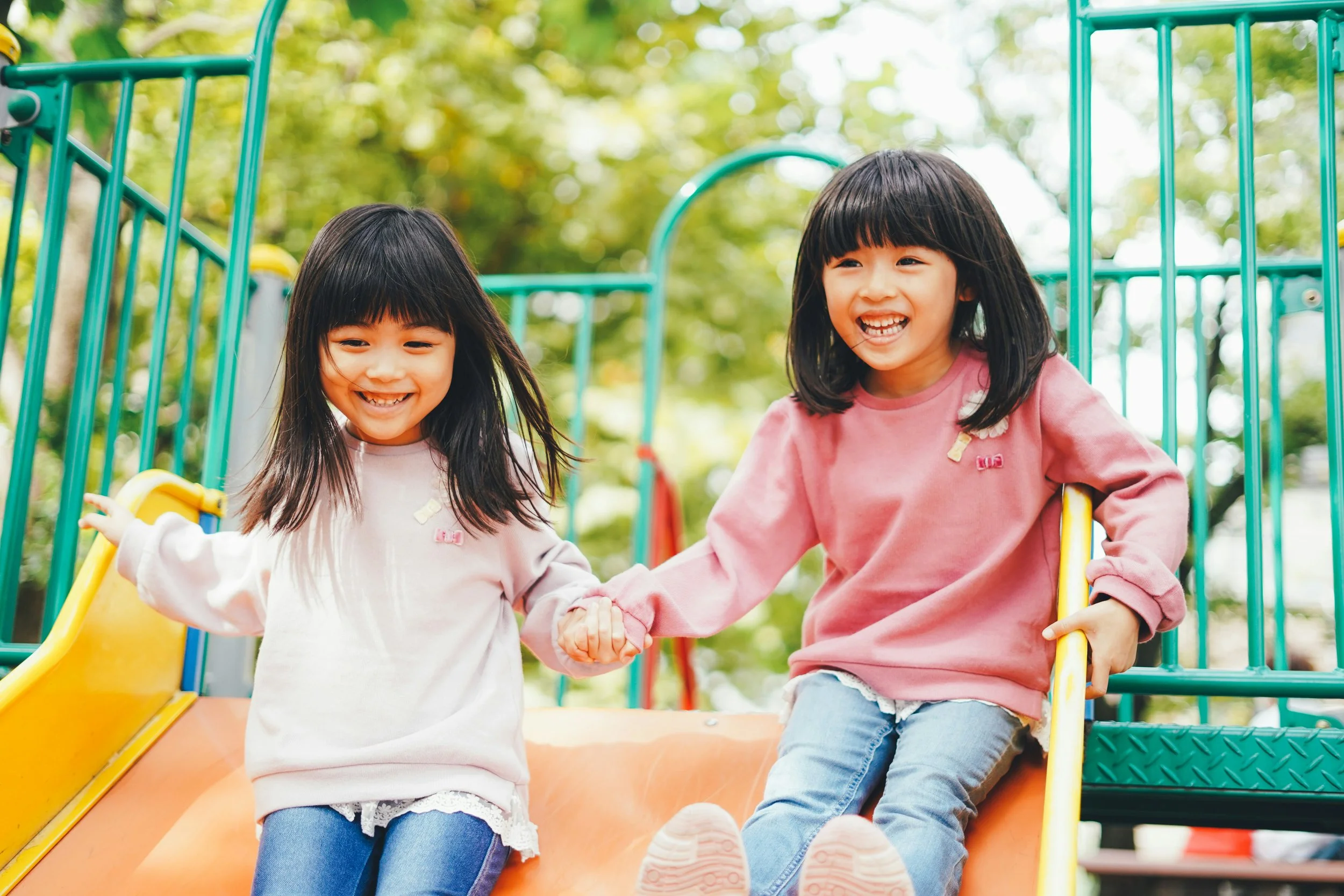 Two young girls with black hair, one with straight hair and the other with wavy hair, are smiling and holding hands while sitting on a slide at a playground. They are outdoors with a blurred green background of trees.
