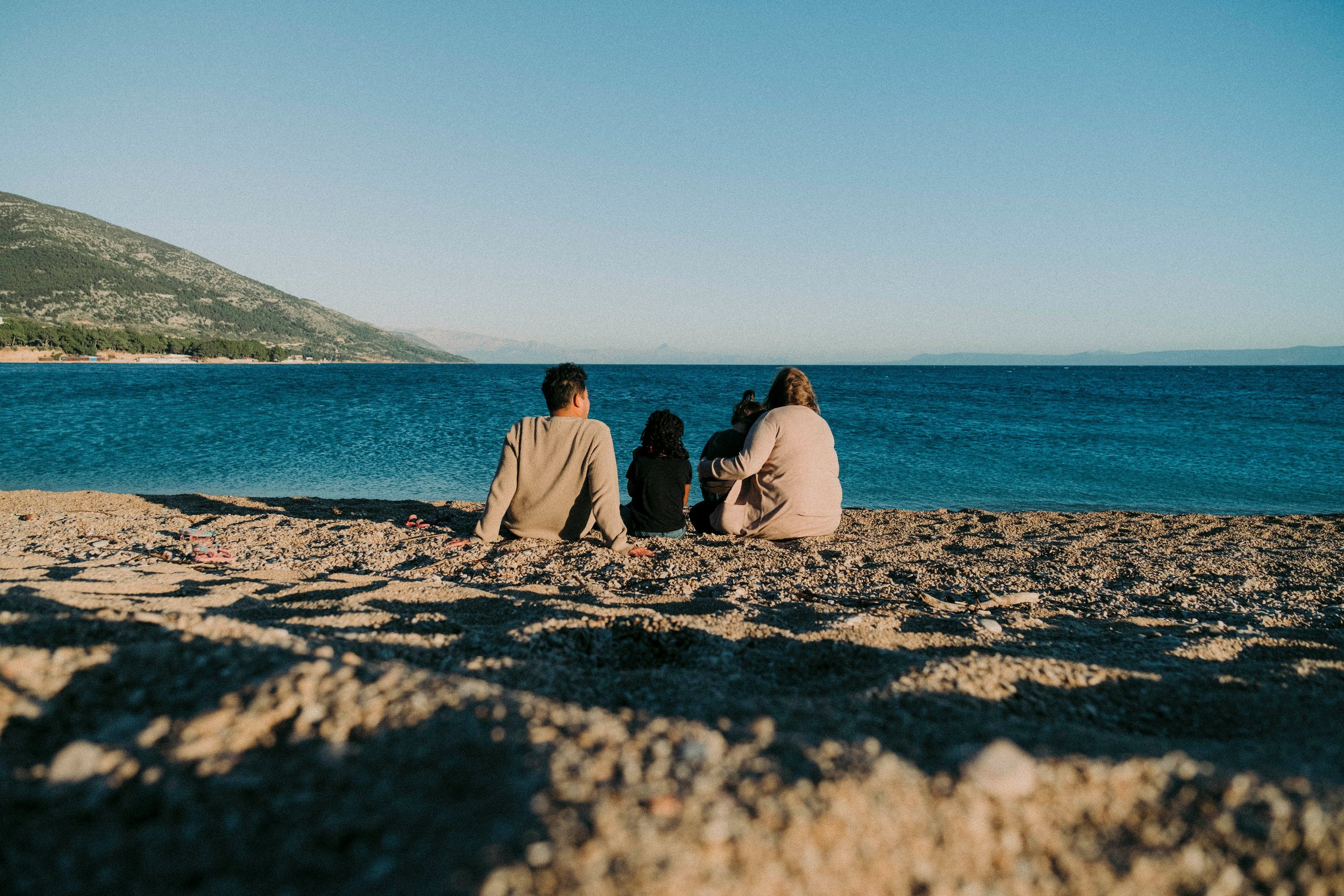 Group of four people sitting on a sandy beach facing the water with mountains in the background.