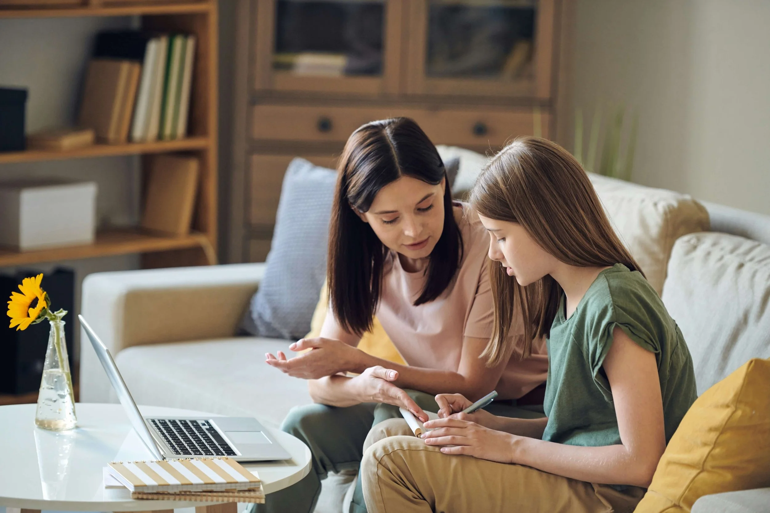 A woman and a young girl sitting on a white sofa, looking at a smartphone together in a cozy living room. There is a laptop, a notebook, and a sunflower in a glass vase on the table in front of them.