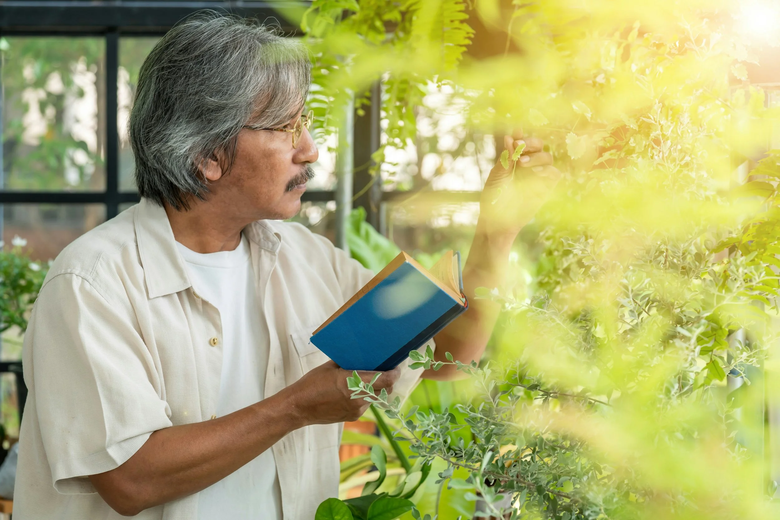 A man with gray hair, glasses, and facial hair examining plants in a greenhouse, holding a blue book.