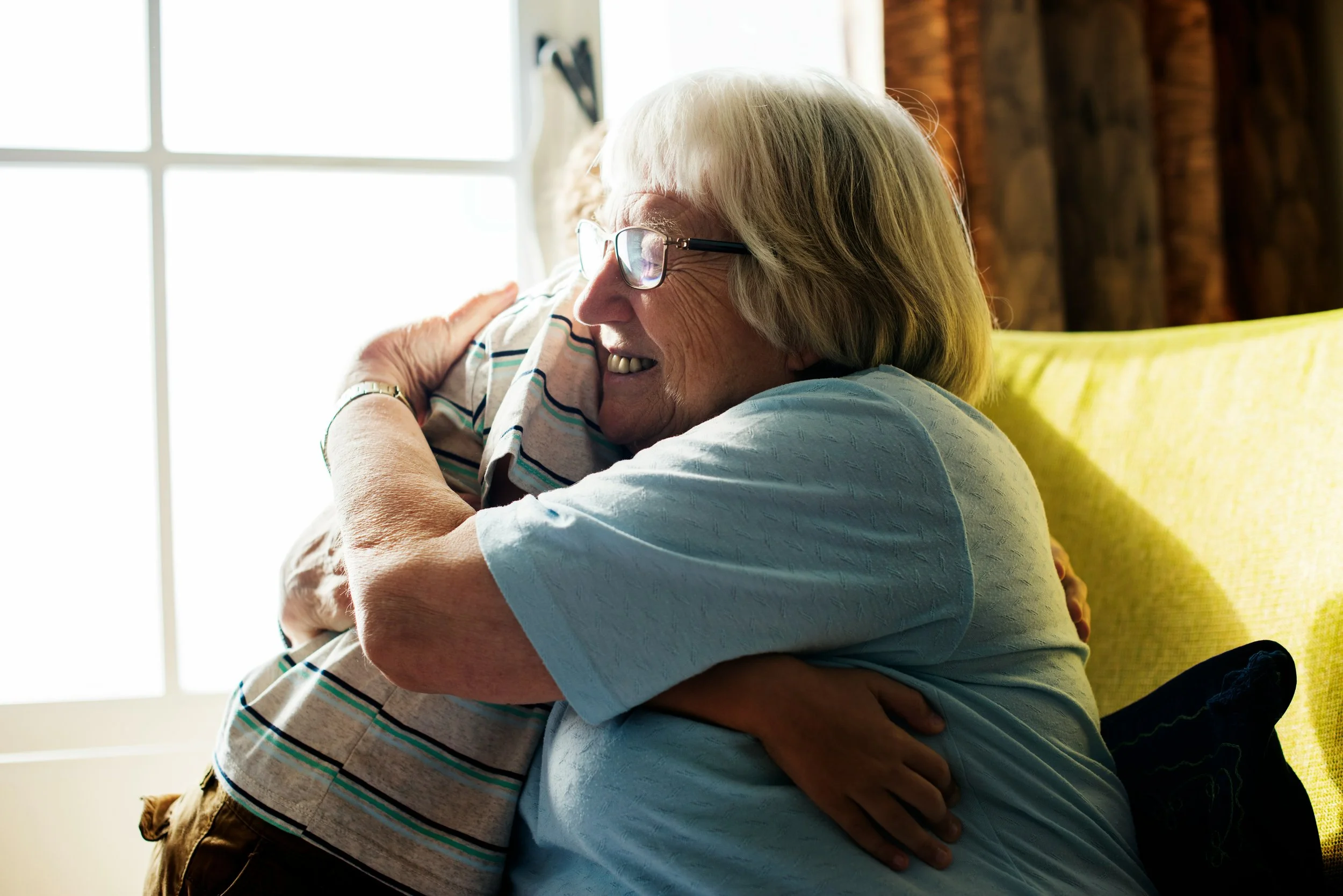 An elderly woman with glasses hugging a young boy inside a house, with sunlight coming through a window in the background.