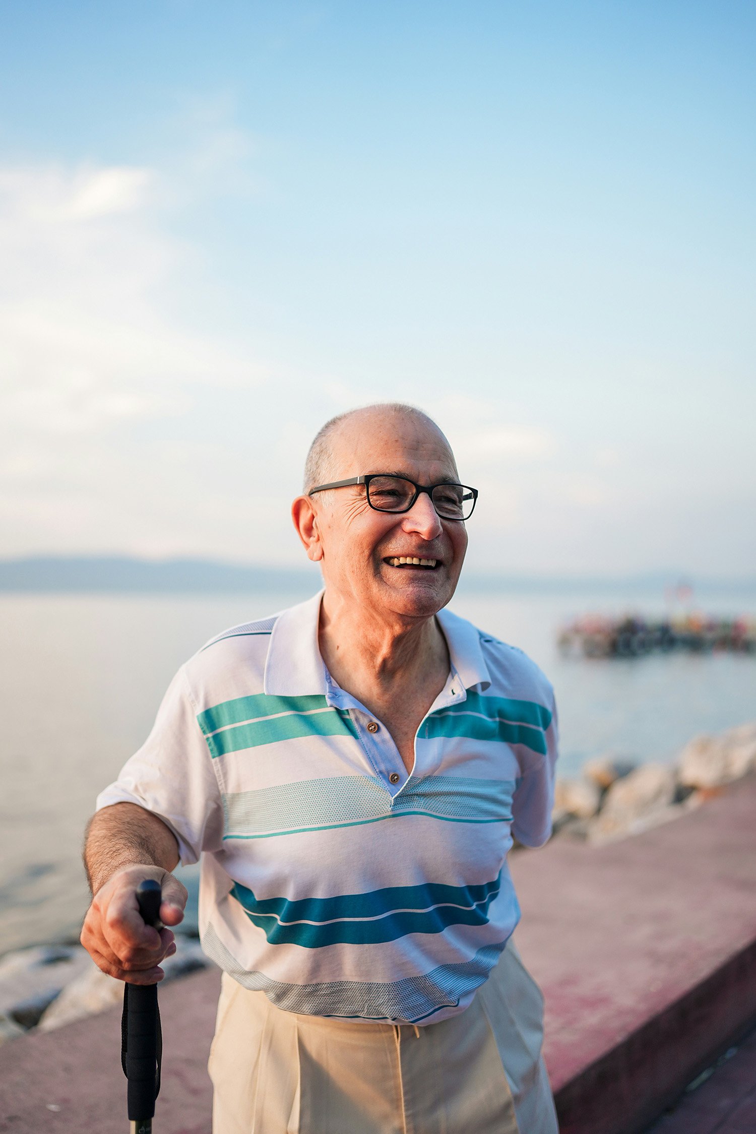 Smiling older man with glasses taking a selfie while walking along a waterfront path during sunset, wearing a striped polo shirt and beige pants, holding a cane in his right hand.