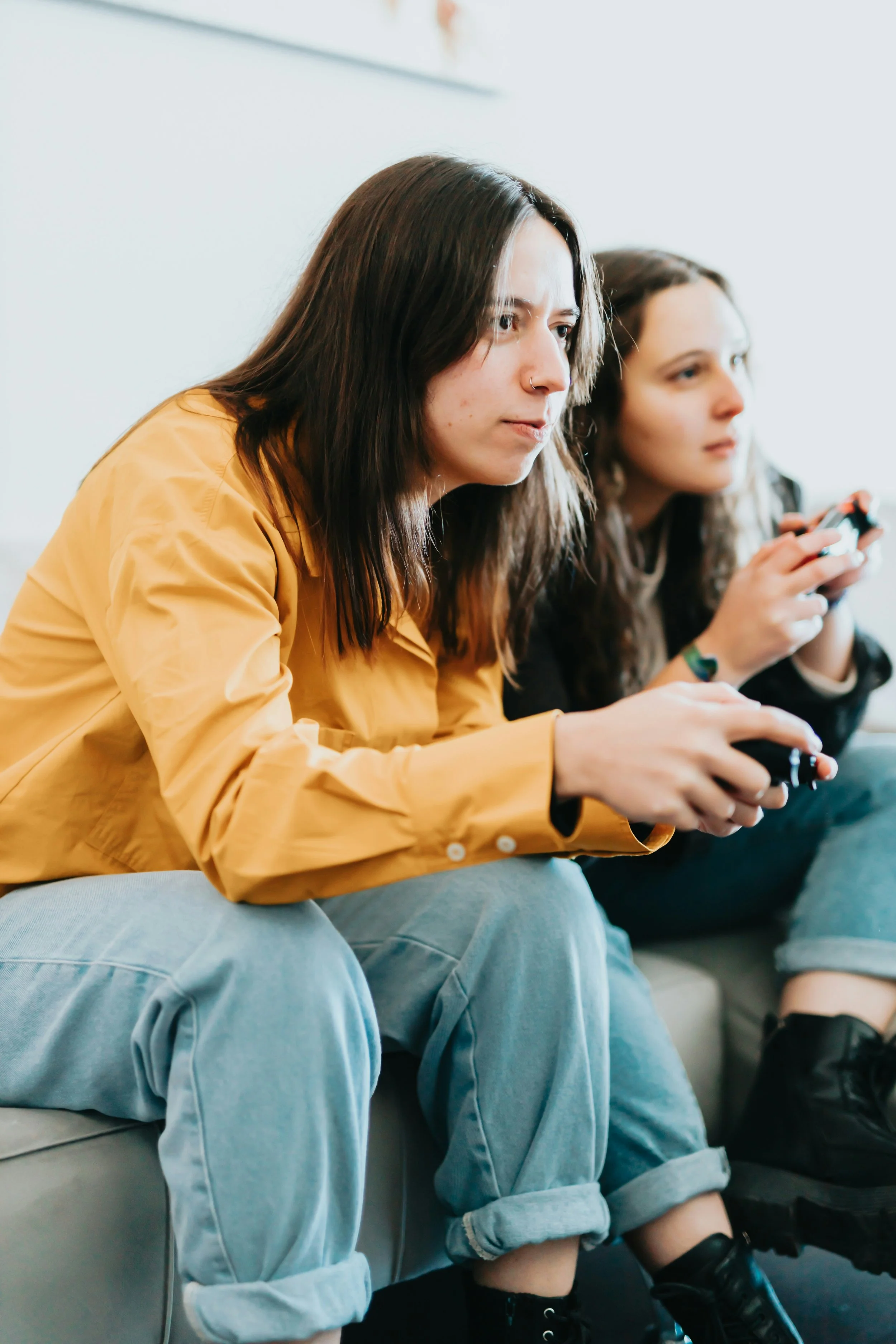 Two young women sitting on a sofa, each engaged with their phones, one wearing a tan jacket and gray jeans, the other in darker clothing, both with long brown hair.