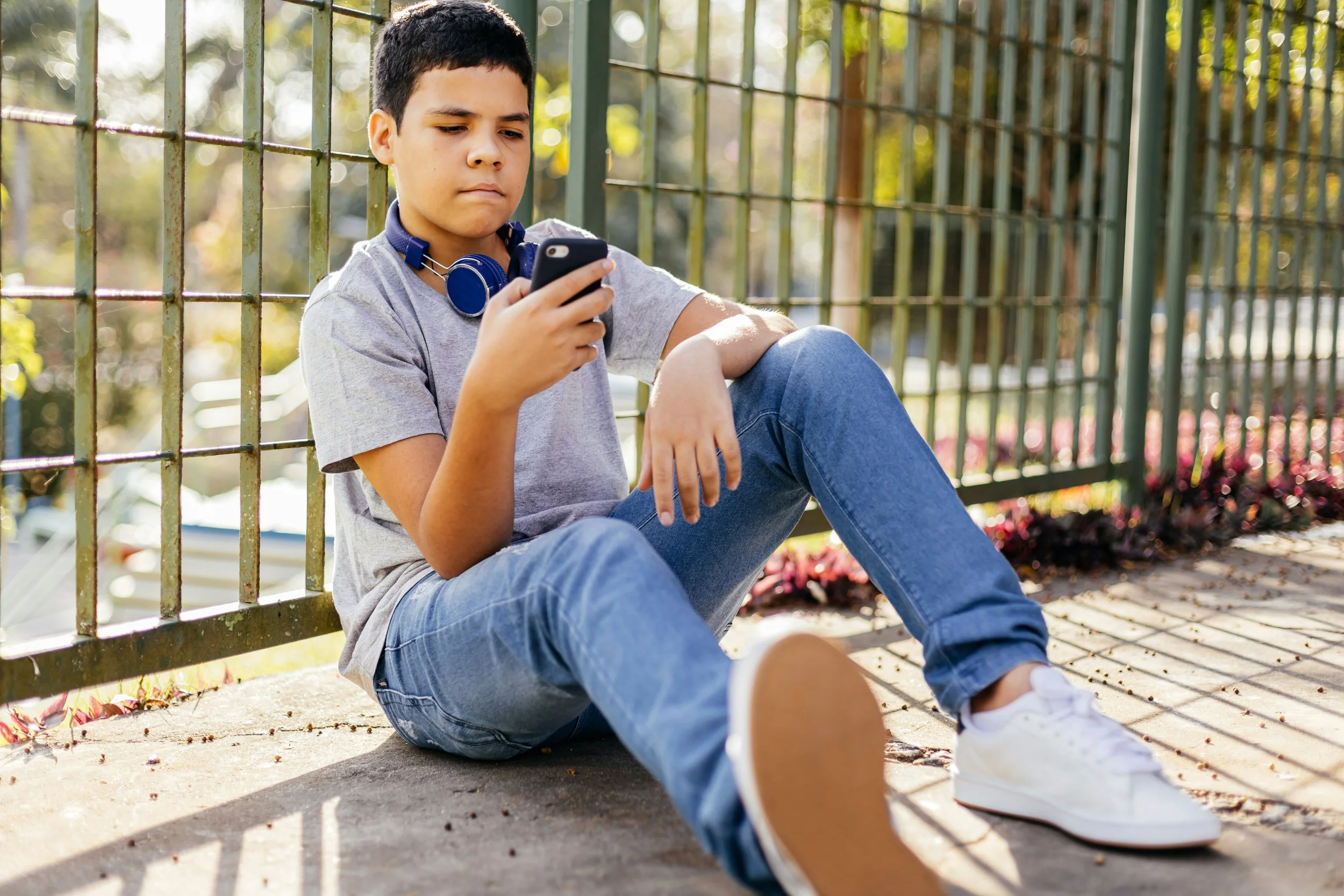 A teenage boy with dark hair sitting on the ground next to a metal fence, looking at his smartphone. He is wearing a gray t-shirt, blue jeans, white sneakers, and has blue headphones around his neck. There are blurred trees and flowers in the background.
