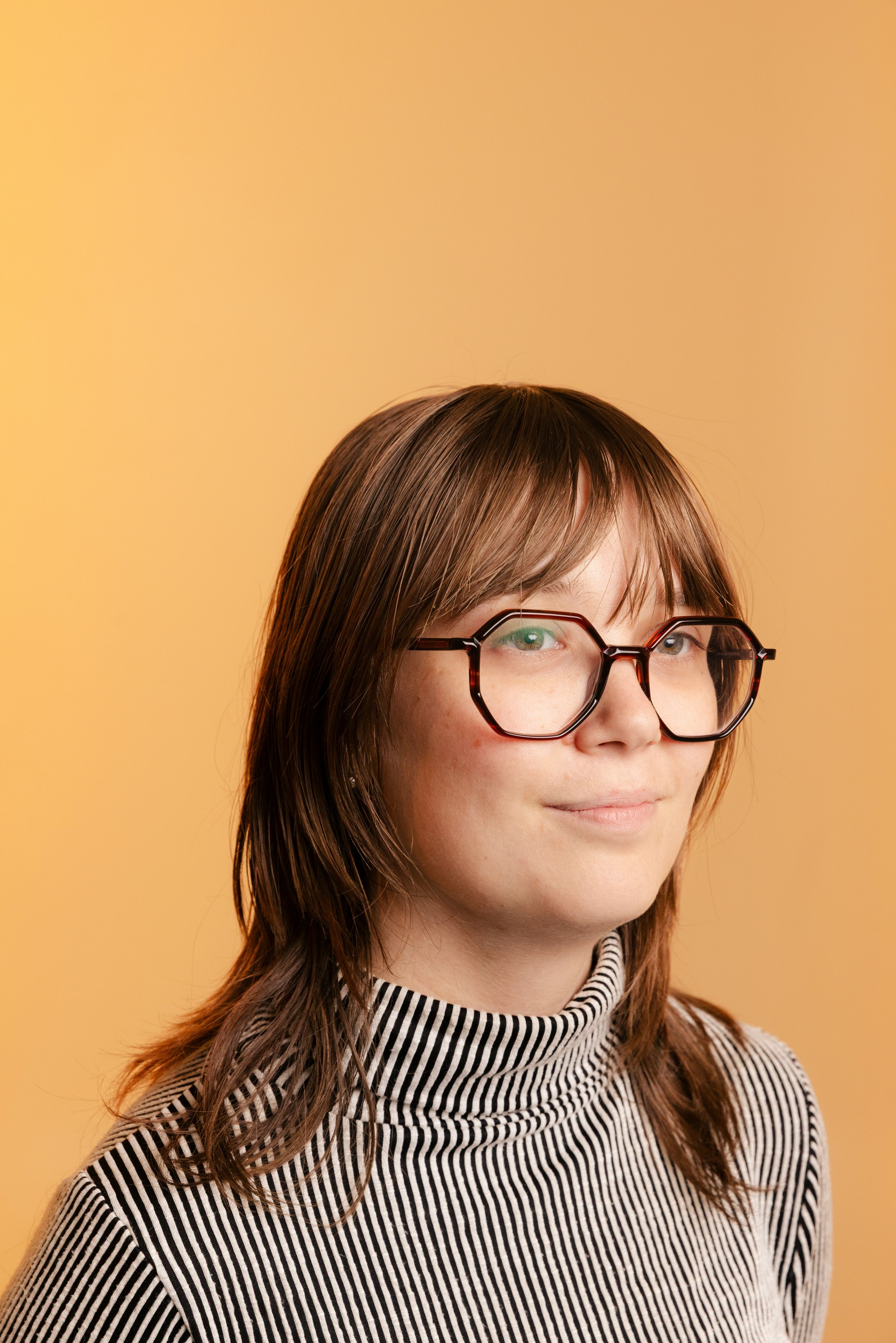 Close-up of a woman with brown hair, wearing black-rimmed glasses and a striped turtleneck, standing against a light orange background.