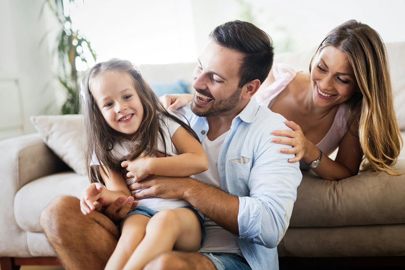 A family of three, a man, woman, and young girl, enjoying a playful moment on a beige sofa in a bright living room.