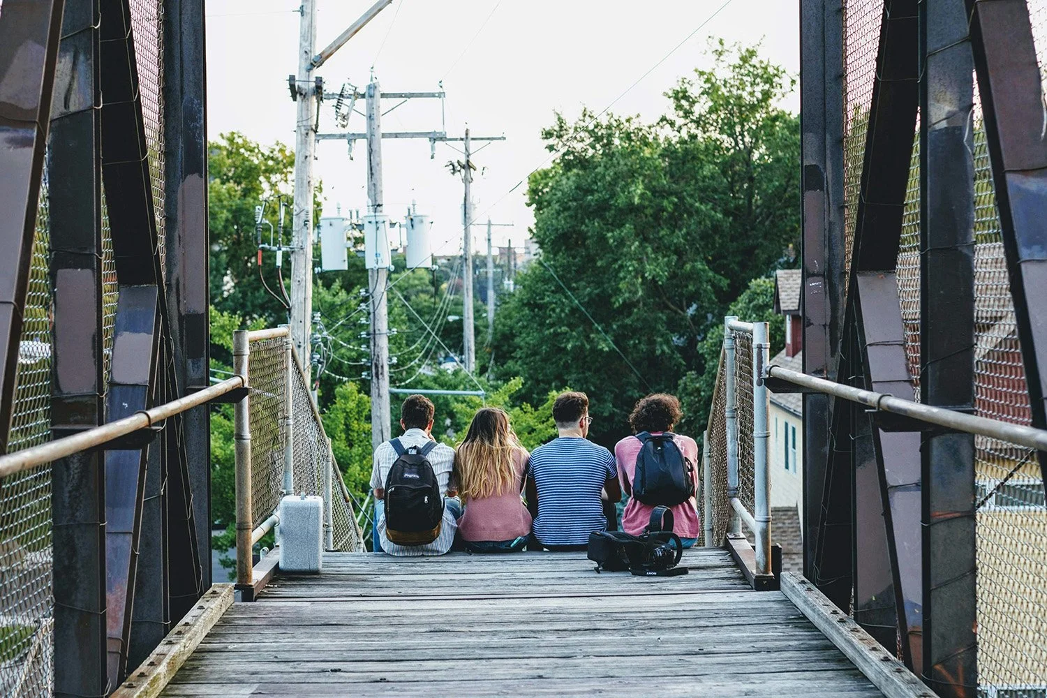 Four young people sitting on a bridge with backpacks, facing away, overlooking a green, tree-filled area and power lines.