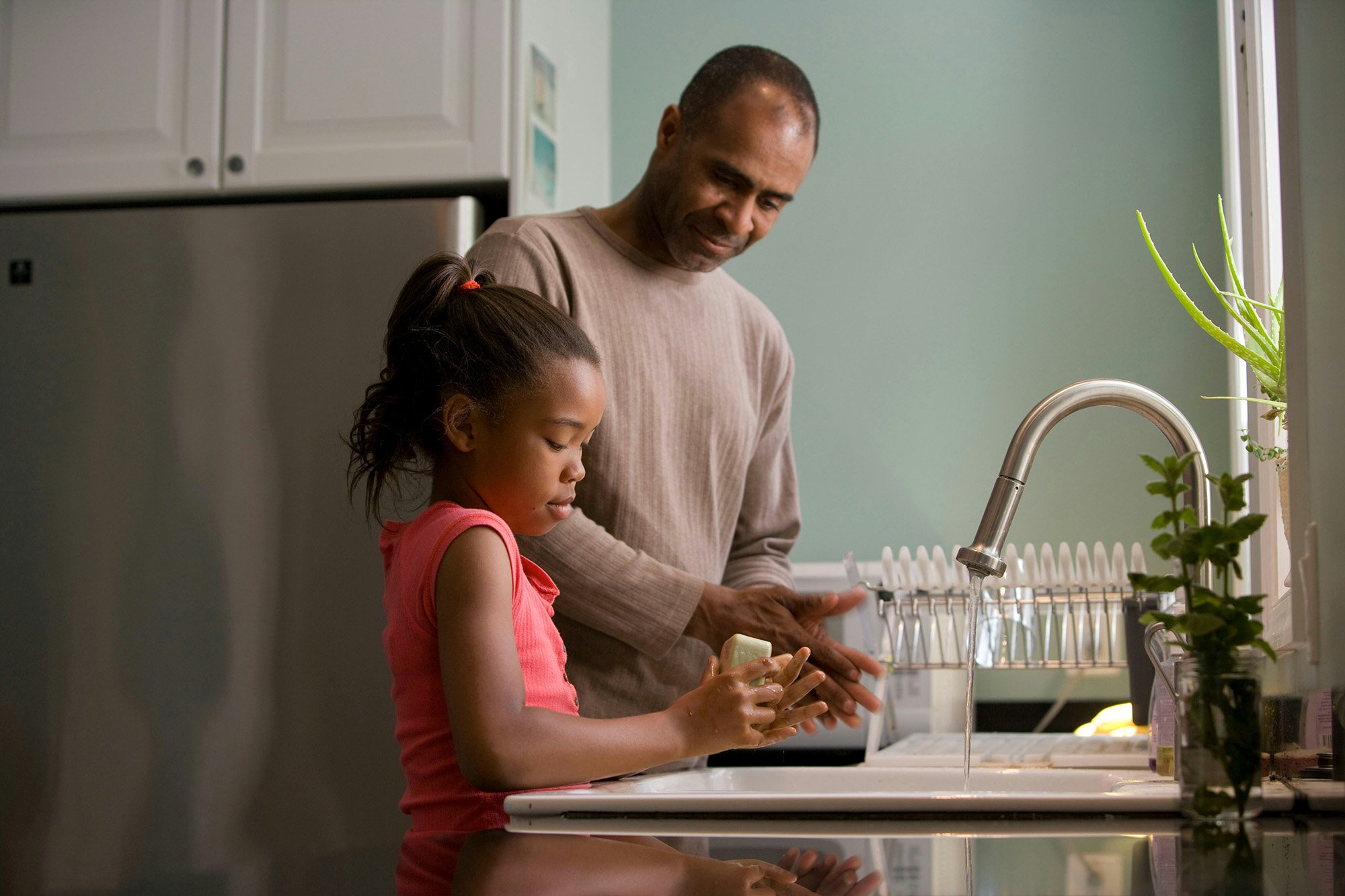 A man and a young girl washing dishes together at a kitchen sink, with greenery visible outside the window.