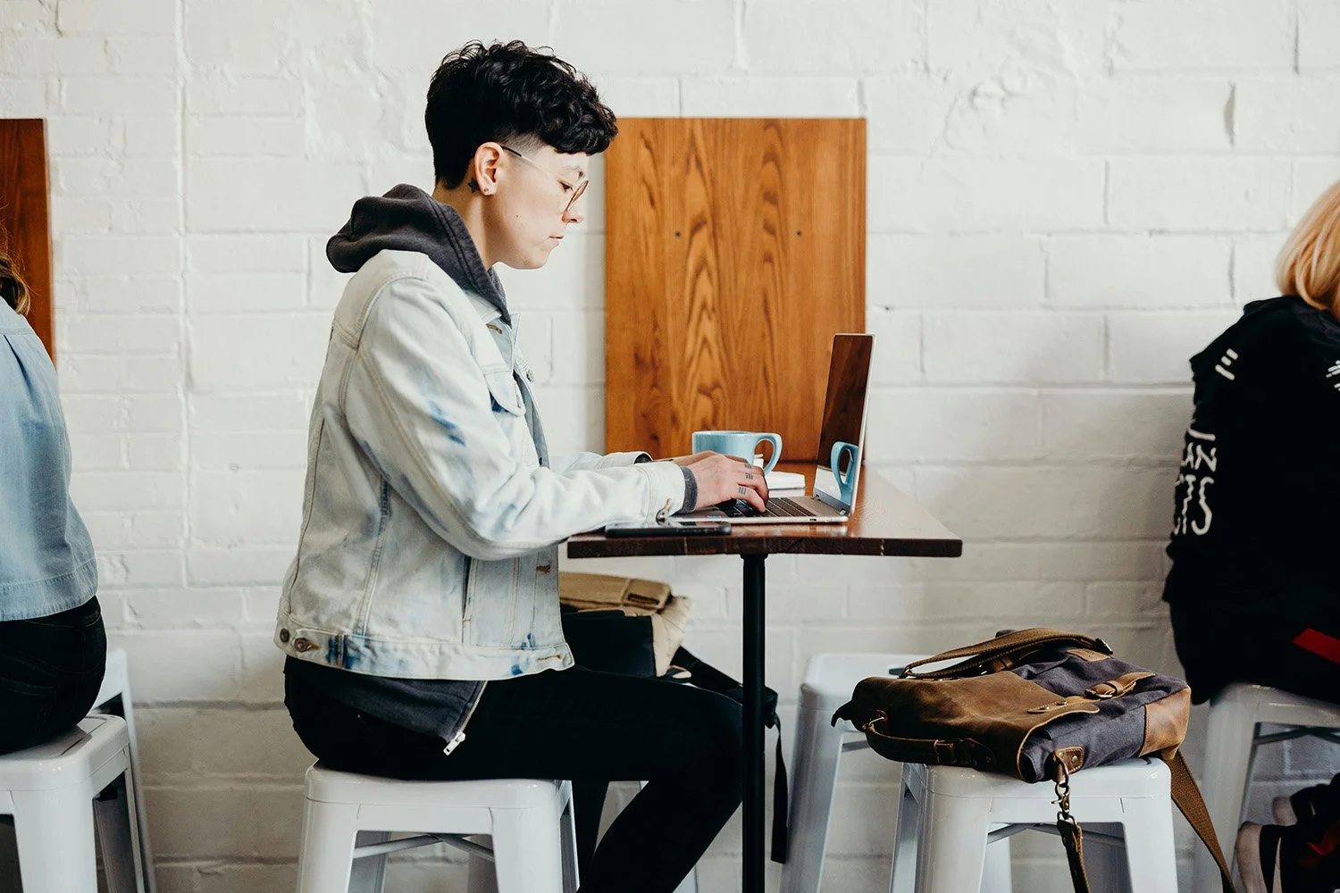 A person sittings on a stool at a tall table, typing on their laptop