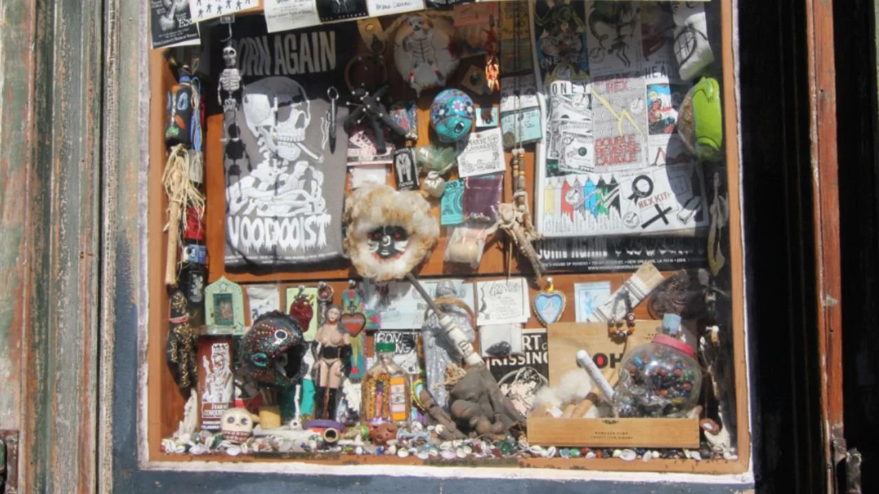 A shop window in New Orleans displaying eclectic voodoo-themed art, skulls, dolls, posters, and colorful trinkets.